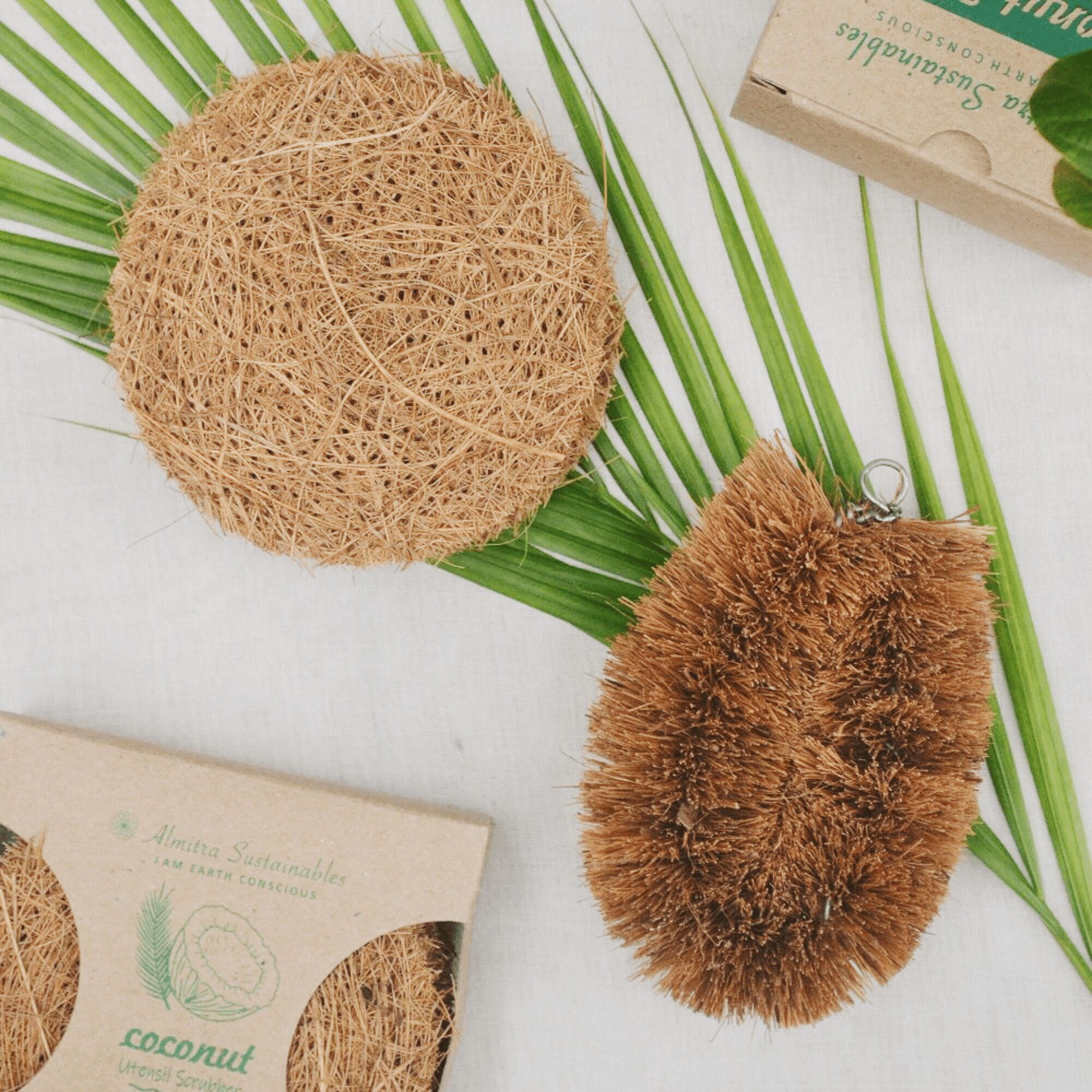 A flat lay of an eco-friendly kitchen cleaning set featuring a round coconut coir scrubber and a vegetable brush made from natural coconut fibers, placed on palm leaves with green foliage and sustainable packaging, reflecting Almitra Sustainables’ commitment to zero-waste living.
