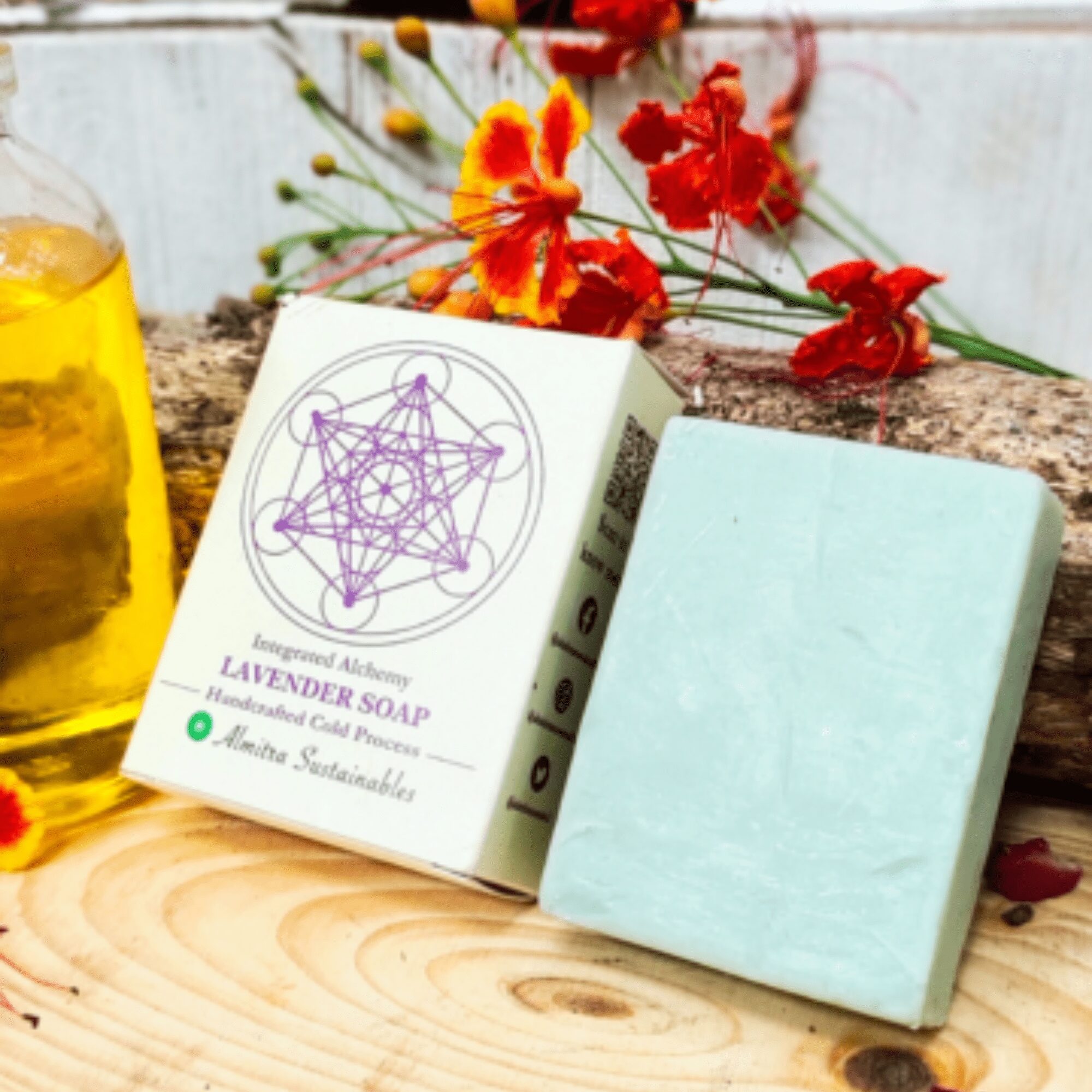 A soothing handmade lavender soap bar by Almitra Sustainables, shown resting on a rustic wooden plank beside its eco-friendly packaging featuring purple geometric design. Surrounded by vibrant red and orange blossoms and a bottle of golden herbal oil, the cold process soap bar reflects a natural, plastic-free skincare choice for eco-conscious users.