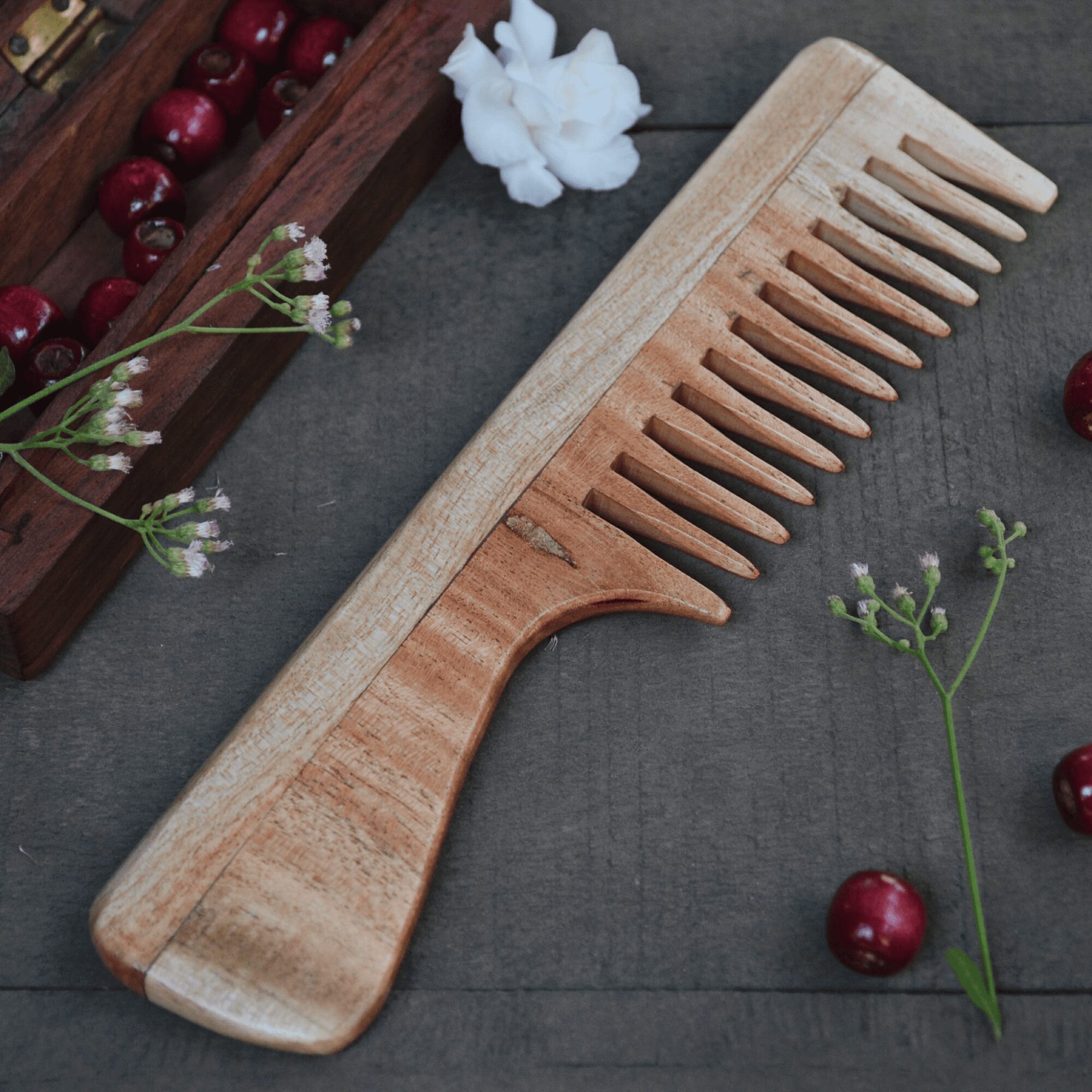 Sustainable wide-tooth neem wood comb with handle placed on rustic wood with cherries and flowers – ideal for detangling thick hair.