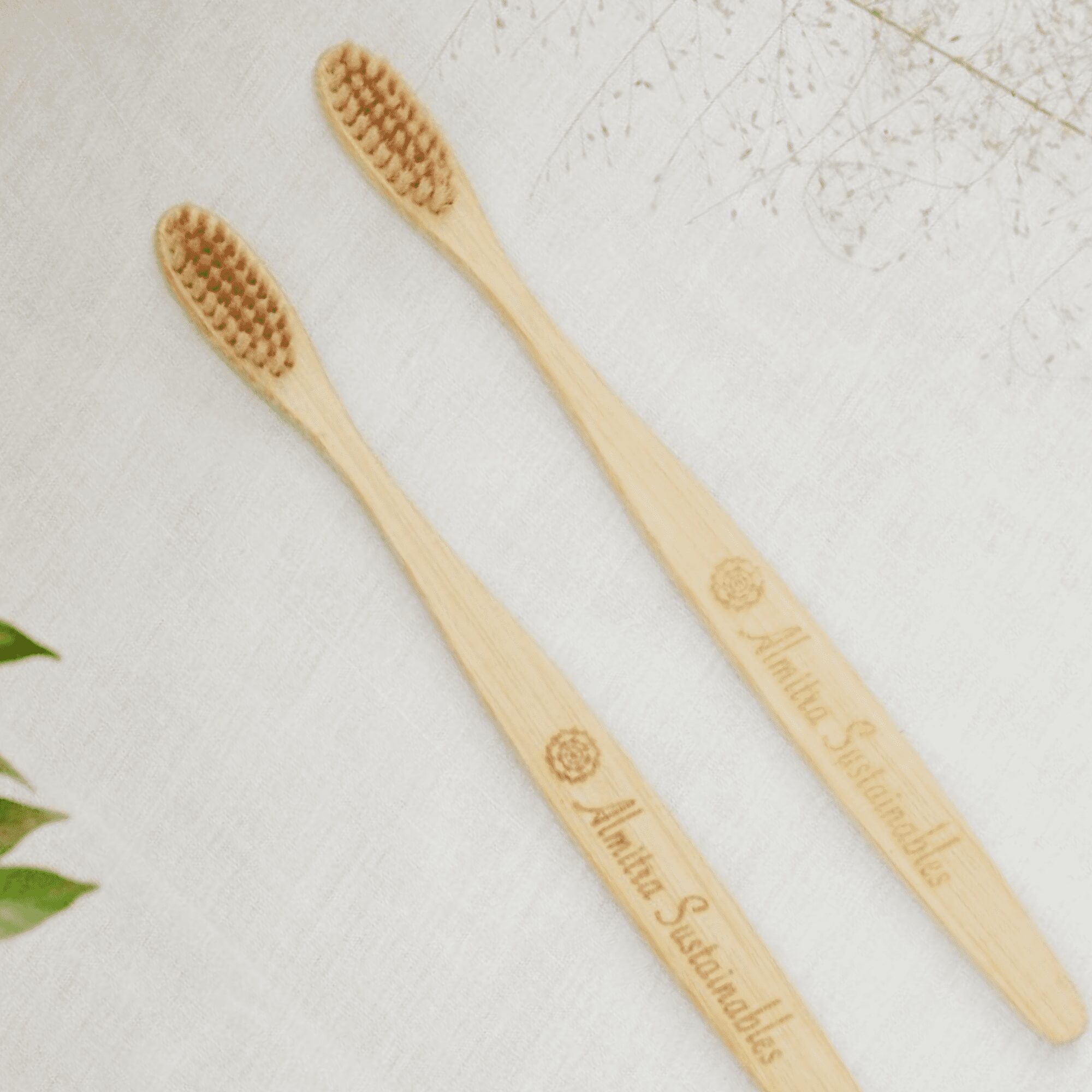 Two sustainable bamboo toothbrushes for kids with soft bristles and “Almitra Sustainables” engraved handles, placed on a fabric surface with greenery.