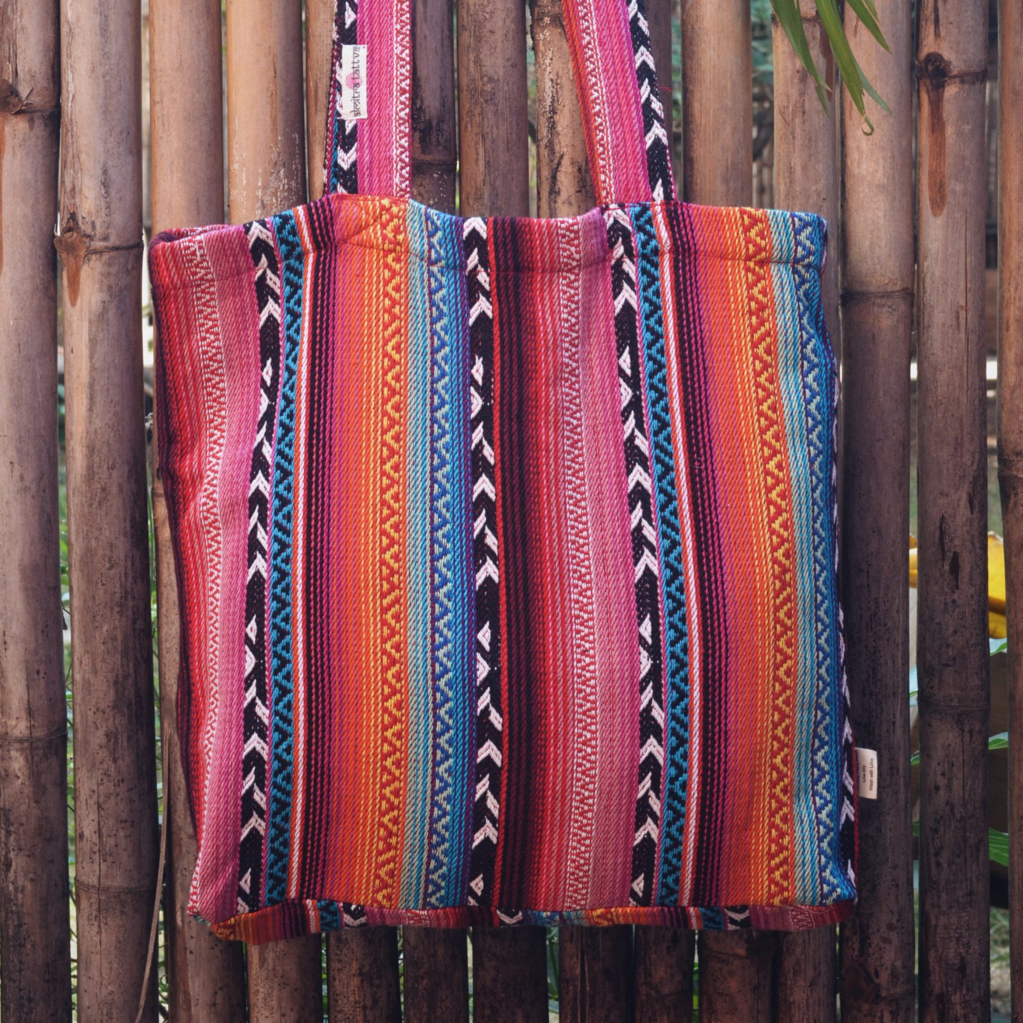 “Larona Tote Bag by Almitra Sustainables, made from vibrant handwoven cotton with traditional multicolor ethnic stripes and geometric patterns, featuring a zip closure and sturdy shoulder straps, sustainably crafted and photographed against a bamboo fence with tropical greenery”