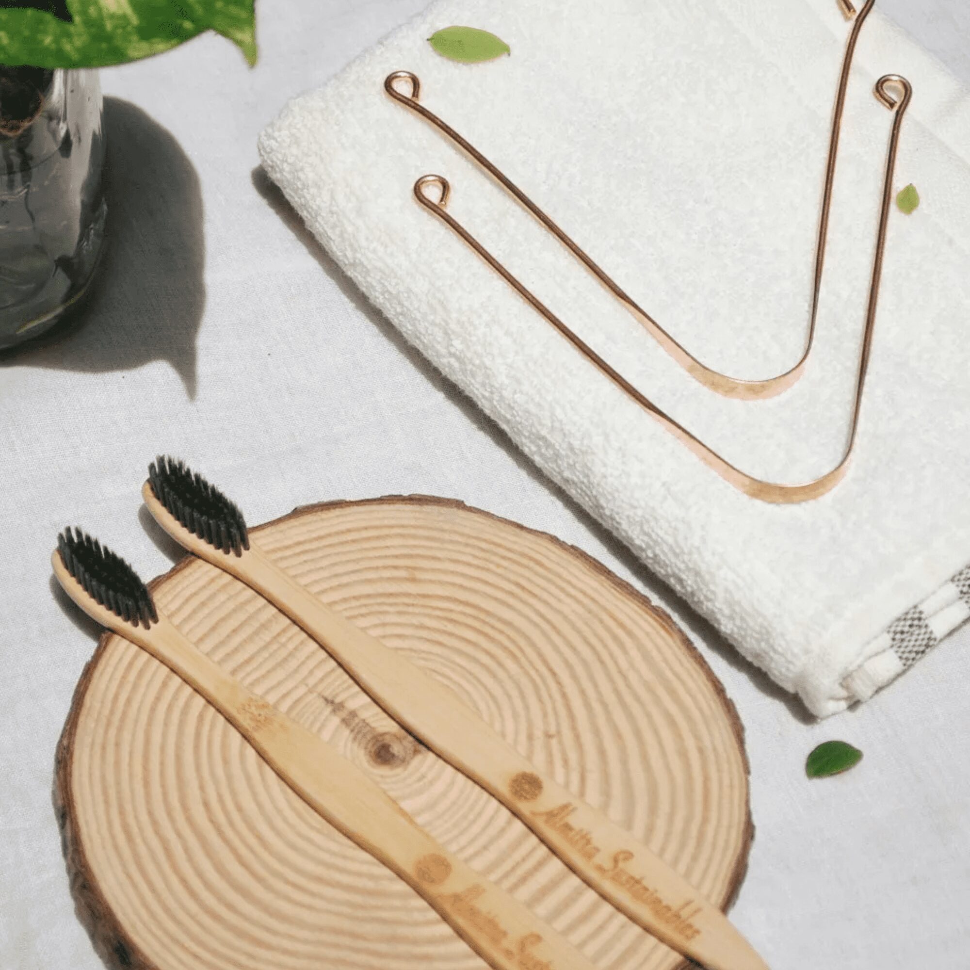 Plastic-free oral care set with two charcoal bamboo toothbrushes and two copper tongue cleaners on a natural wooden slab with a white towel"