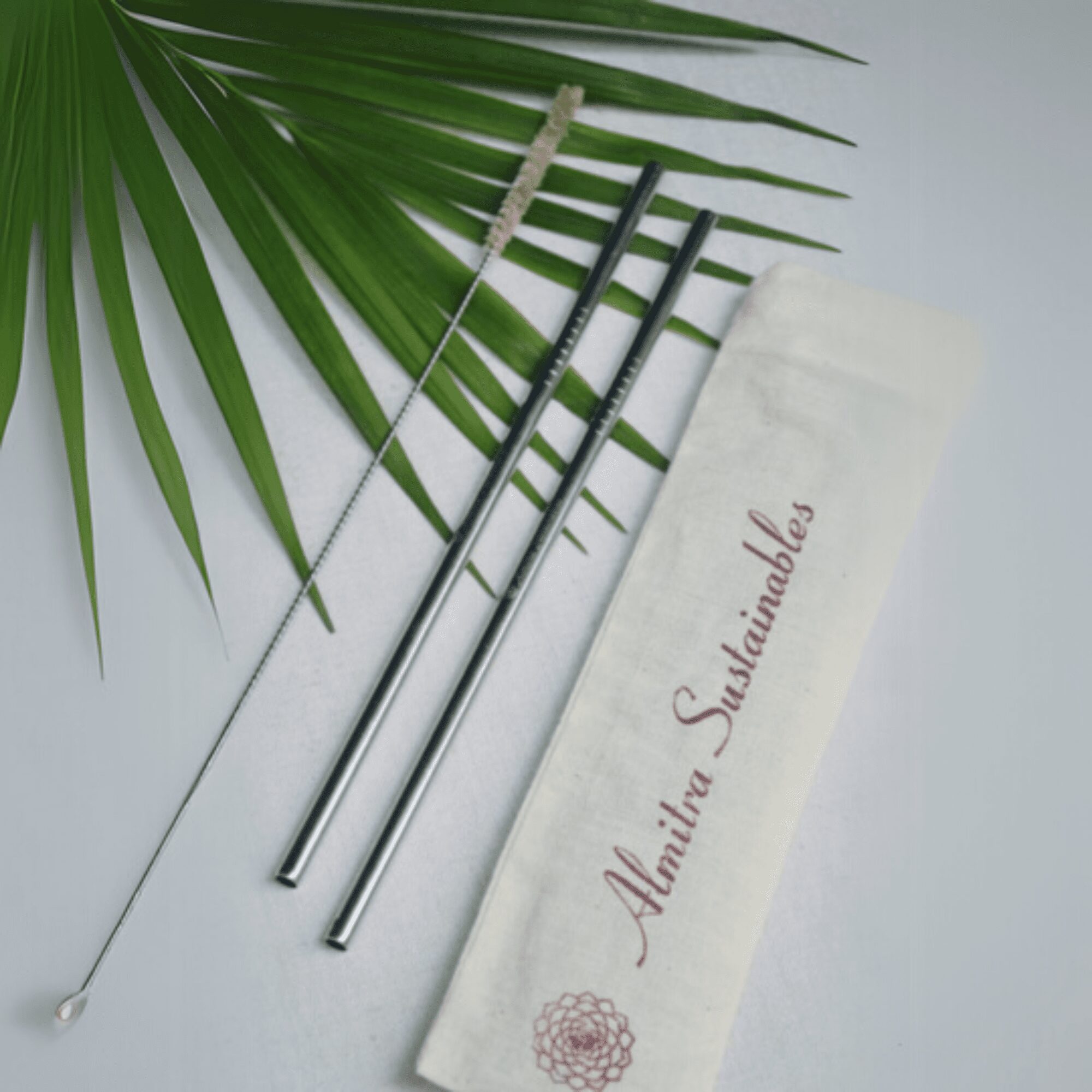 Flat lay of a reusable stainless steel straw set by Almitra Sustainables, featuring two eco-friendly metal straws, a straw cleaning brush, and a washable organic cotton pouch, placed over a green palm leaf for a natural, sustainable aesthetic.
